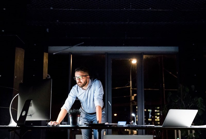 Thoughtful hipster businessman in his office working late at night at desk with computer and laptop