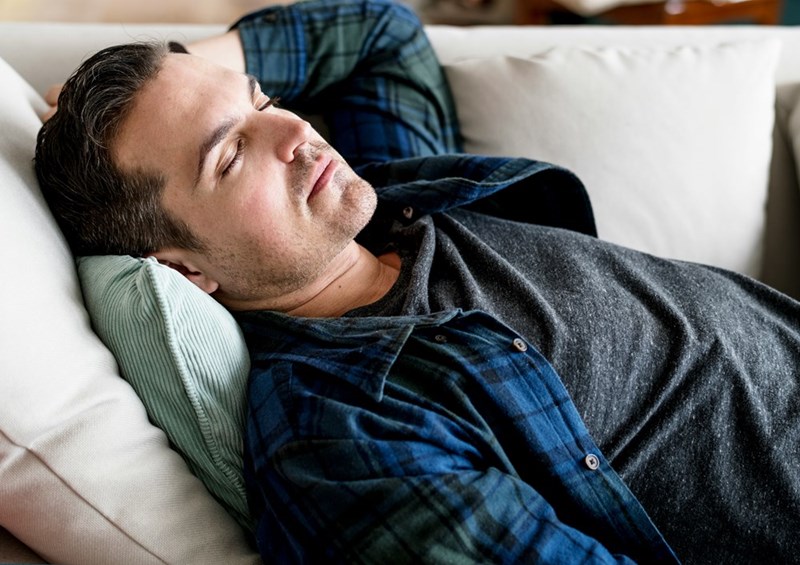 Man resting on a couch with eyes closed and hands behind his head indoors.