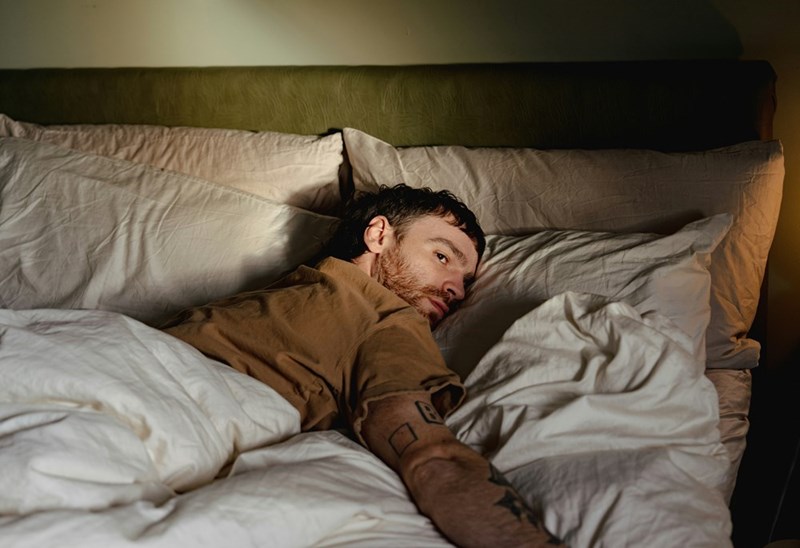 Man lying awake in bed under white blankets in a dimly lit bedroom.