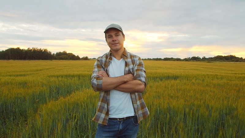 Man in a plaid shirt and cap standing in a grassy field at sunset with arms crossed.