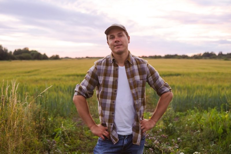 Man in a plaid shirt and cap standing in a grassy field at sunset with hands on hips.
