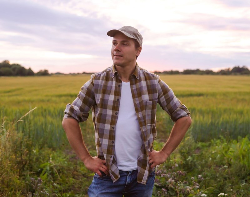 Man in a plaid shirt and cap standing in a grassy field at sunset looking into the distance.