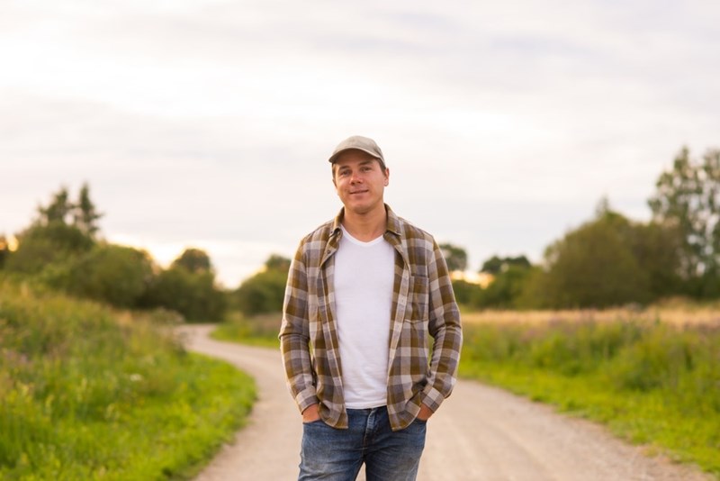Man in a plaid shirt and cap standing on a country road surrounded by grassy fields.
