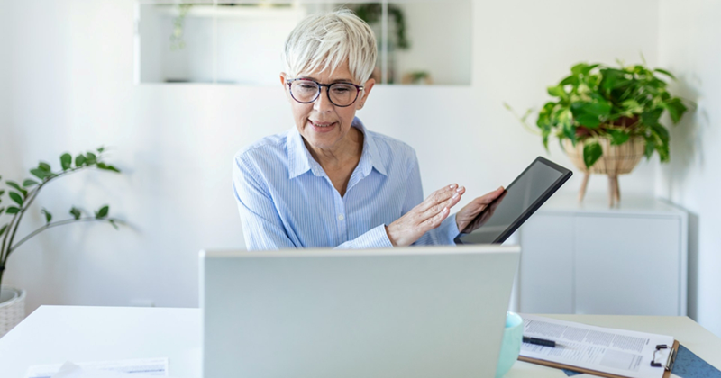 An older employee gestures to a tablet she is holding as she takes a meeting on her laptop at home
