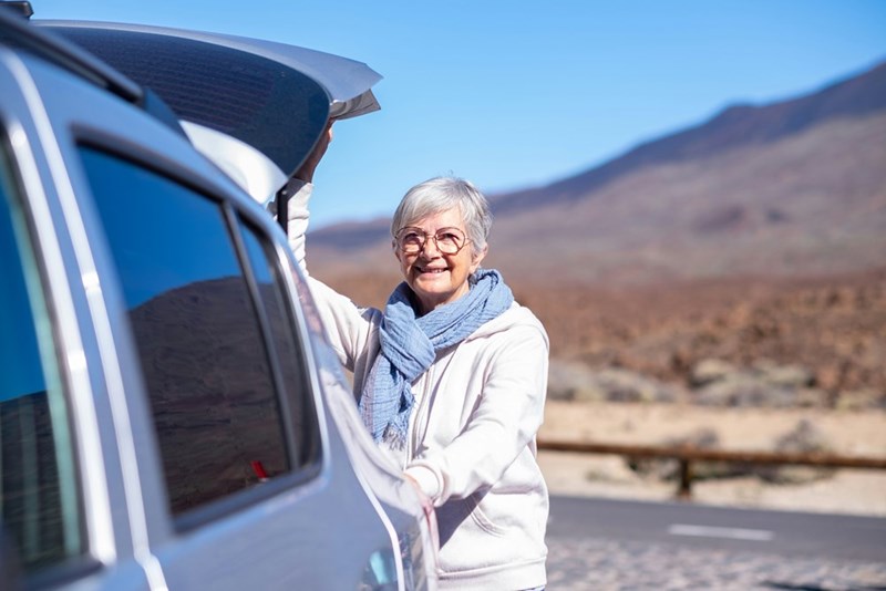 Smiling mature senior woman in outdoors mountain excursion with automobile opening hood of the car