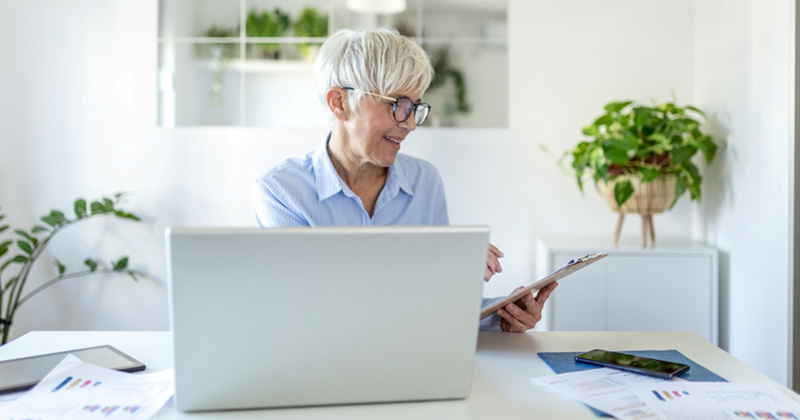 An older employee works on her laptop from home while looking at a clipboard