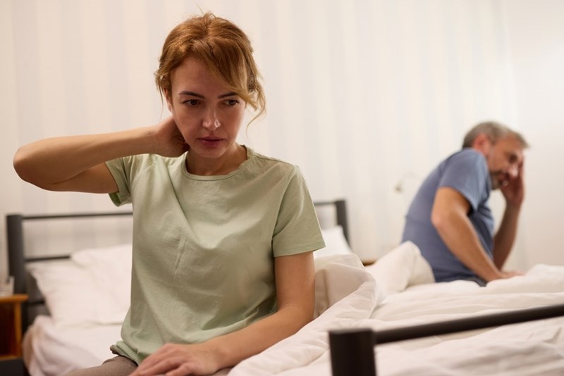 Upset woman sitting on a bed while a man sits in the background in a bedroom.