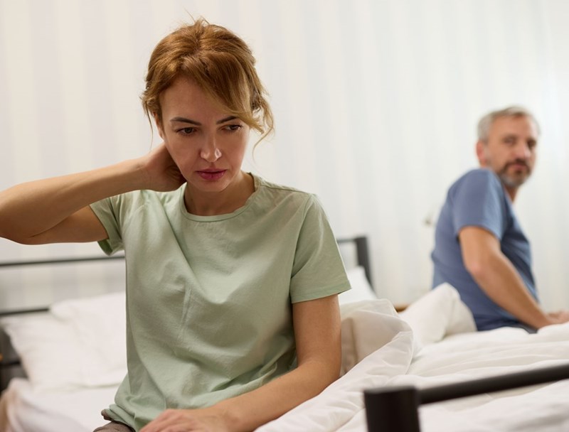 Concerned woman sitting on a bed with a man blurred in the background in a bedroom.