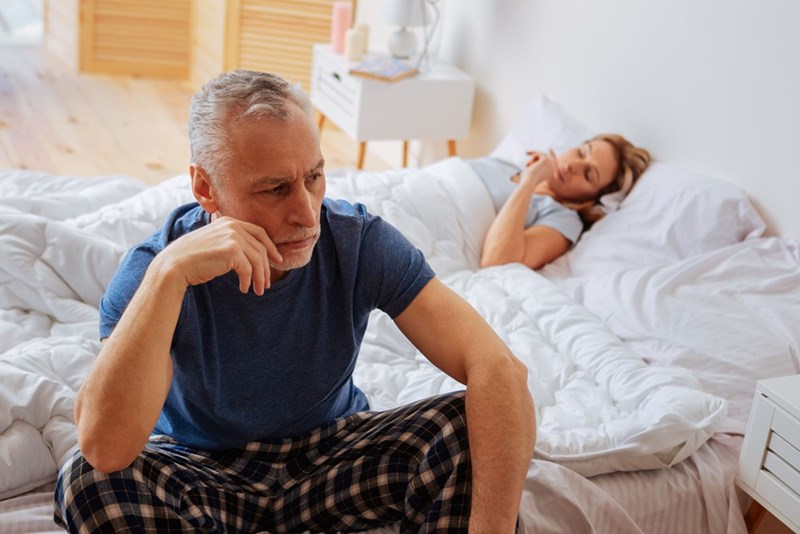 Worried man sitting on a bed while a woman lies awake in the background of a bedroom.