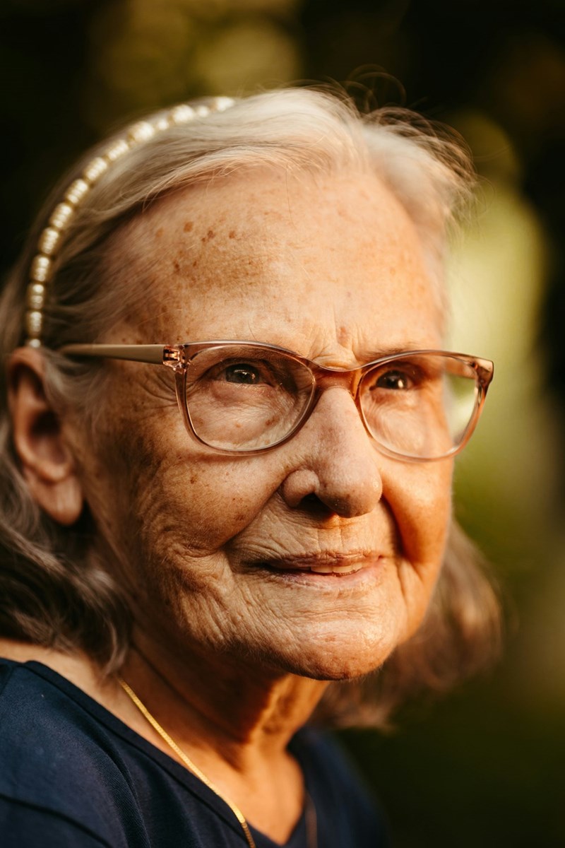 An older woman with glasses wears a pearl headband.