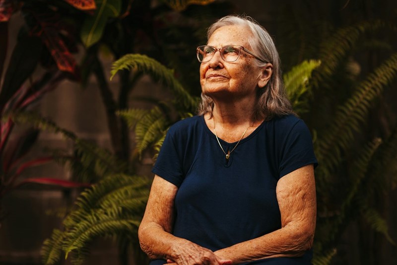 An older woman with glasses looks up as she sits in front of a large plant on her lawn.