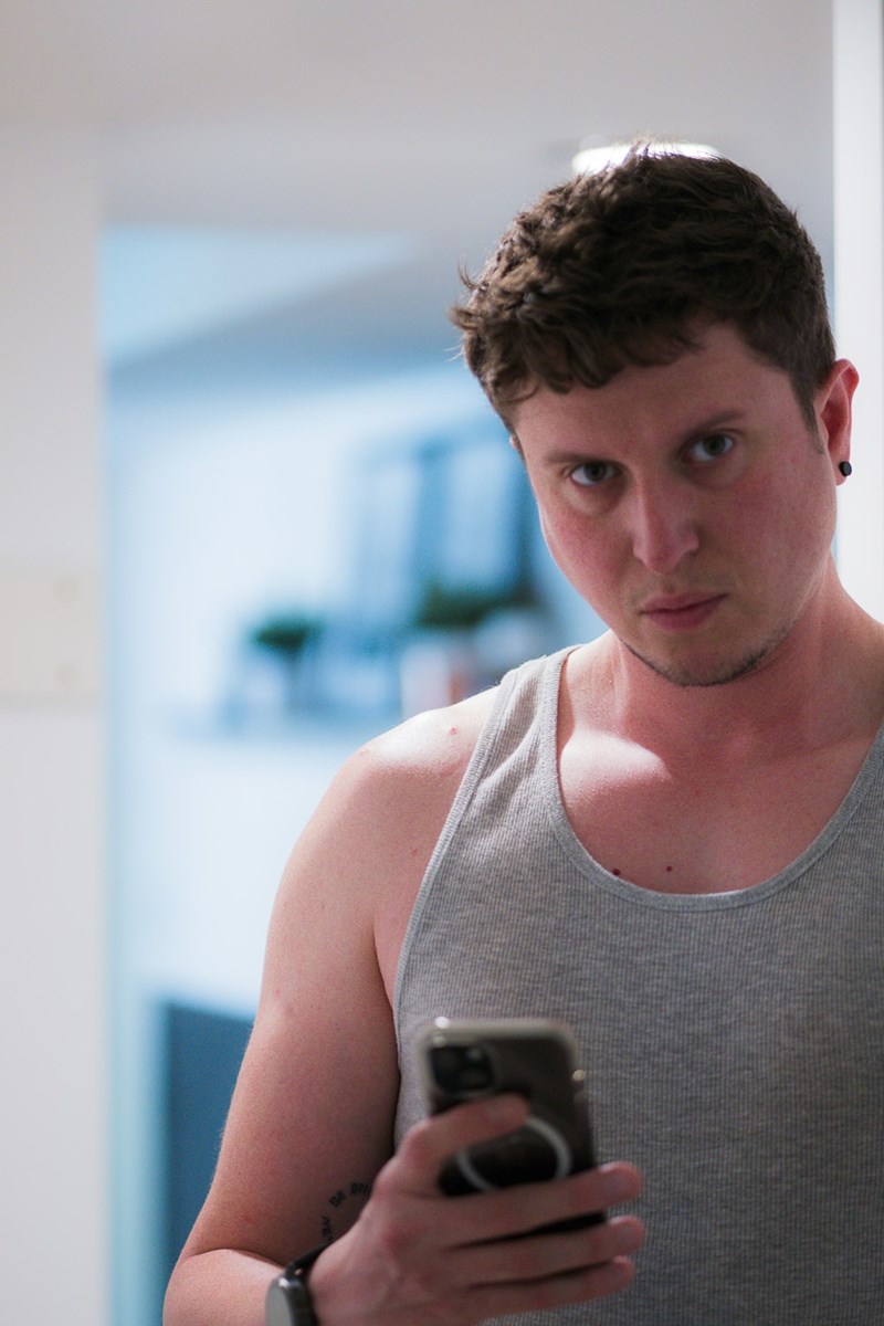 Angry man wearing a grey tank top holding a phone with one hand and looking straight to the camera.