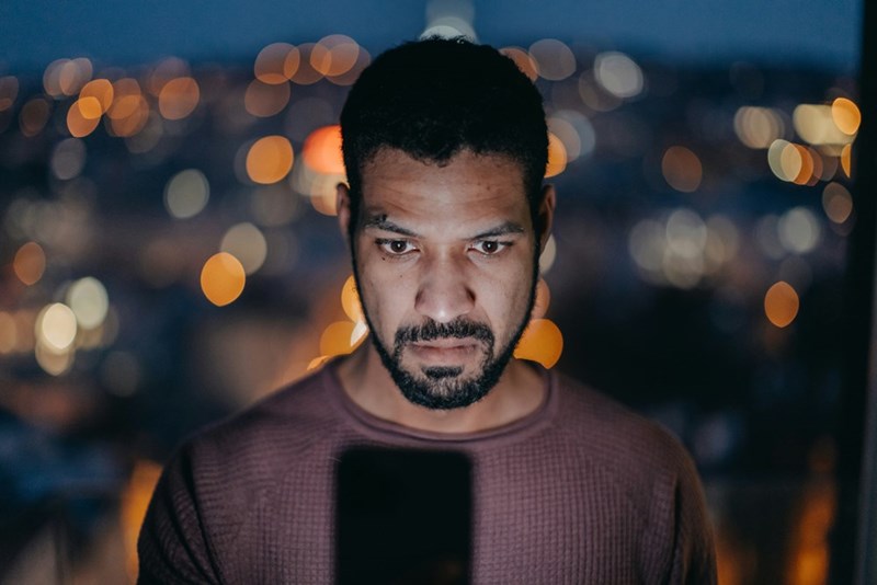 Man with a serious face standing in a high building balcony looking at his phone with city lights behind him.