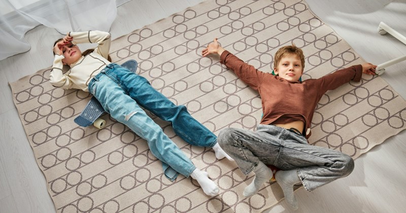A girl lies on a small skateboard and a boy lies with his arms outstretched on top of a rug