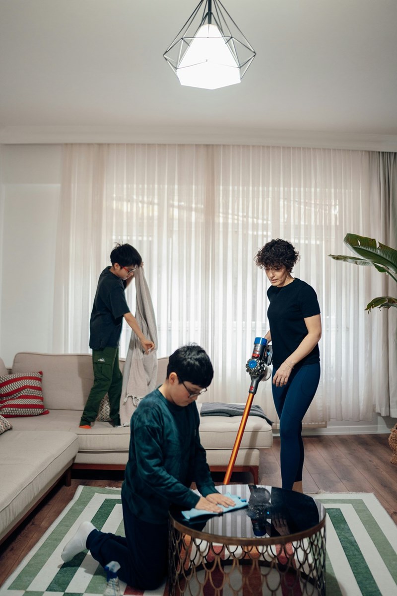 Family cleaning a living room together with kids wiping surfaces and a woman vacuuming the floor.