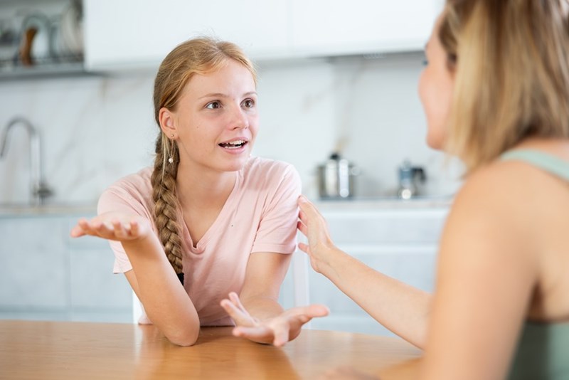 Teen girl speaking with her mother at a kitchen table during a family discussion at home.