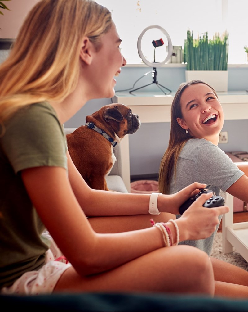 Teen girls playing video games together at home while laughing with their pet dog nearby.