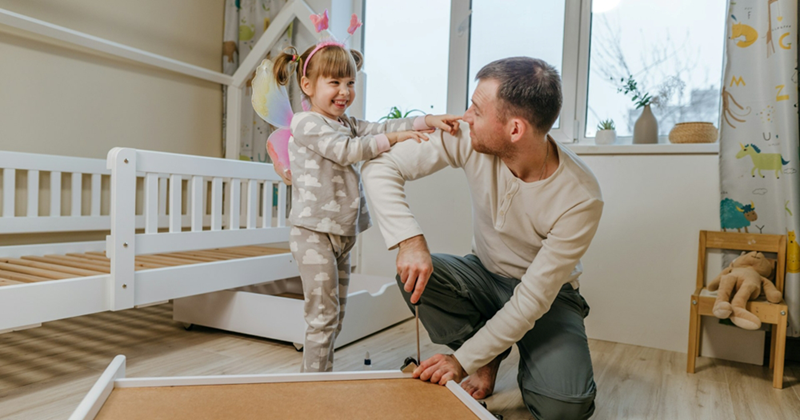 A young girl wearing pink fairy wings smiles and touches the arm of her father, who is assembling a bed in a bedroom