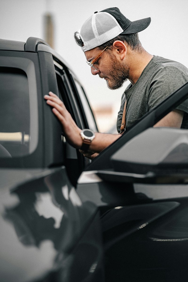 Man in glasses and a baseball cap checking inside a car parked outdoors.
