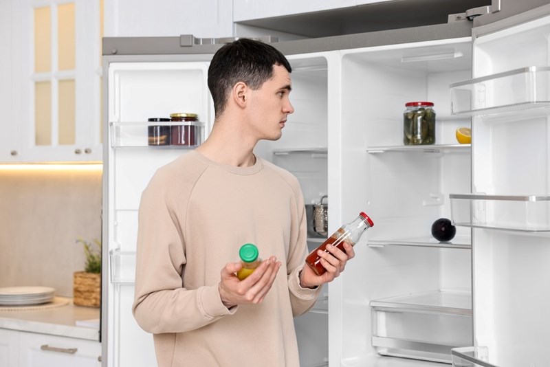 Man holding sauce bottles while checking a nearly empty refrigerator in a modern kitchen.