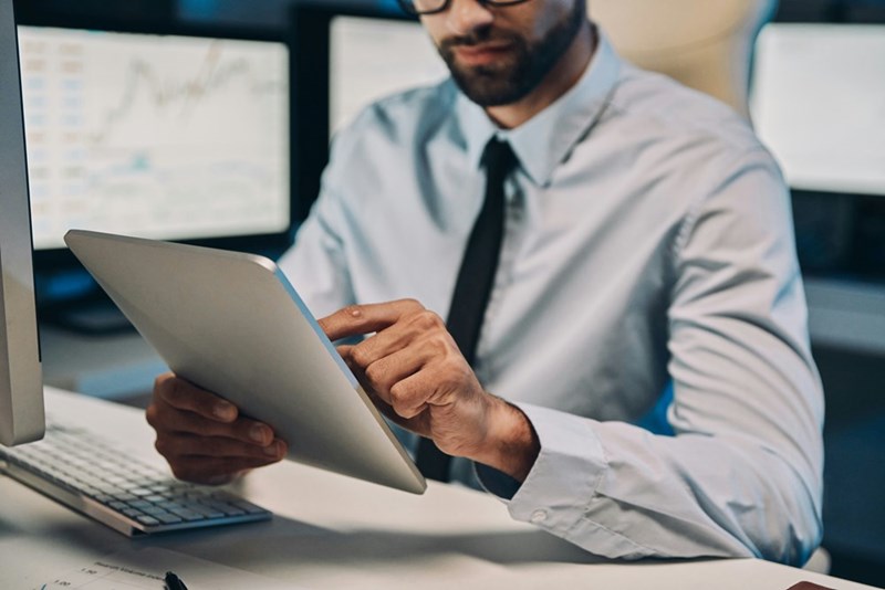 A male employee taps on his tablet while surrounded by computer monitors.