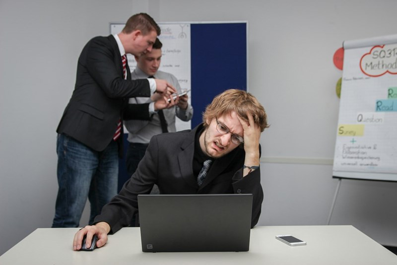 A young male employee leans his head against his hand in front of his laptop as two coworkers discuss something behind him.