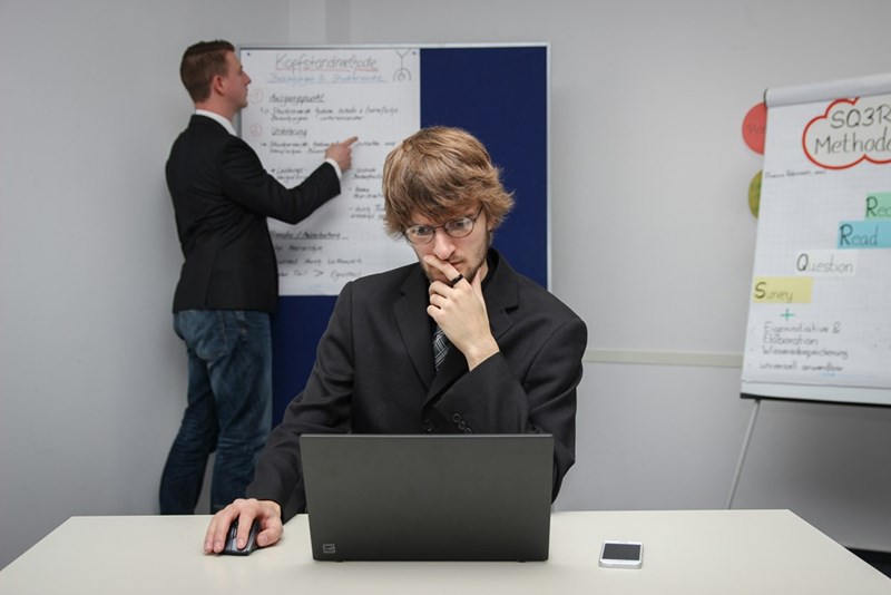 A young male employee looks inquisitive while reading on his laptop as a coworker looks at a whiteboard behind him.