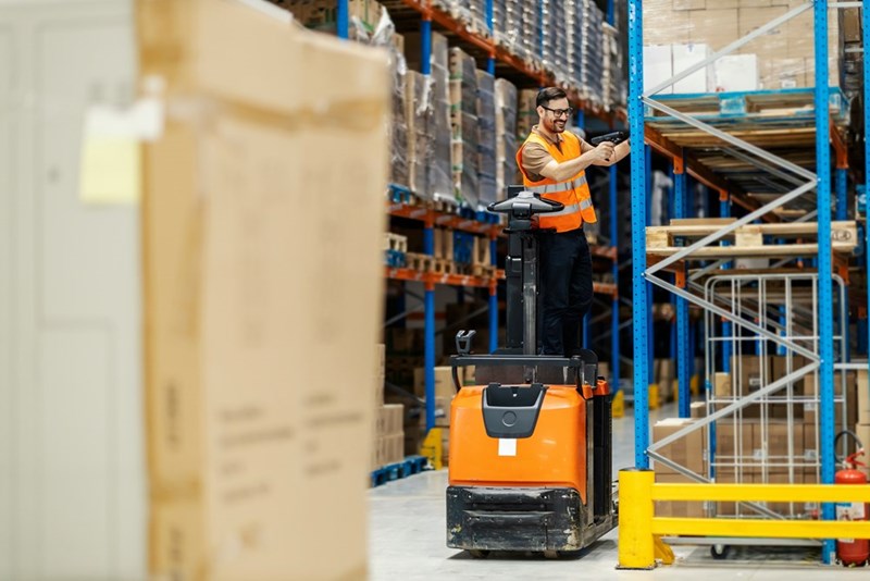 A warehouse employee holds a scanner in his hand while standing on a forklift.
