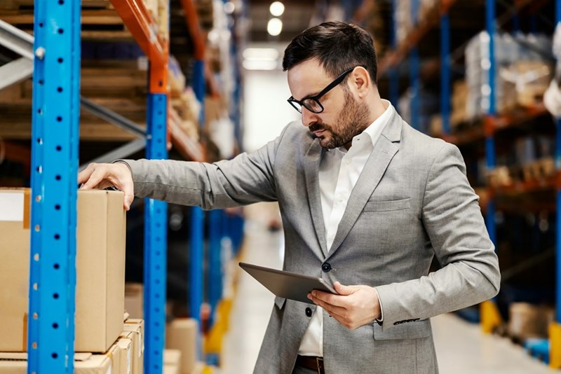 A supervisor wearing a suit checks boxes in storage while holding a tablet.
