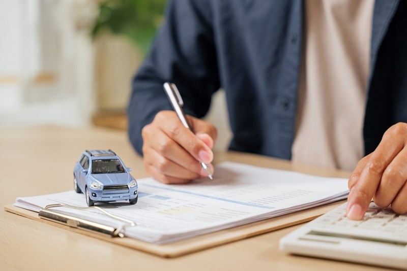 Person calculating car insurance or auto loan costs while signing documents beside a blue model SUV.