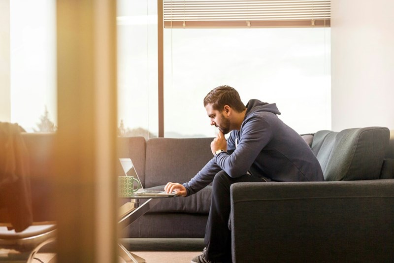 Man working on a laptop from a living room couch while thinking and reviewing information online.