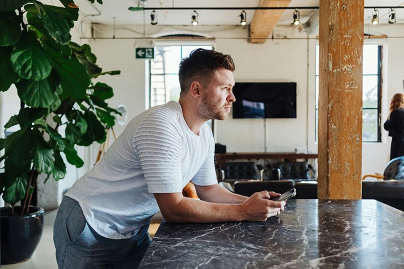Man checking his phone while leaning on a counter in a modern open office with natural light.