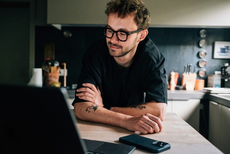 Man wearing glasses working on a laptop at a kitchen counter with a phone beside him.