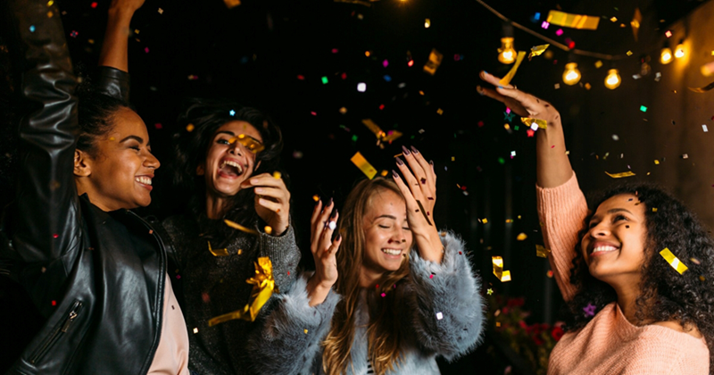 Four women smile and dance with gold confetti falling around them