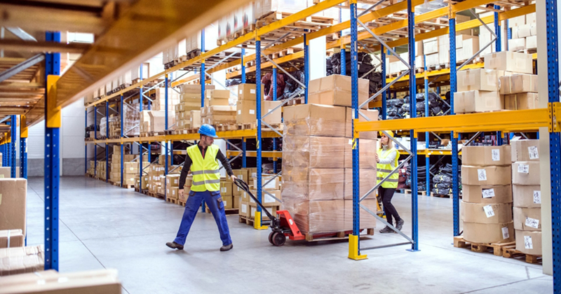 A man in safety gear pulls a pallet through a warehouse with a woman behind the pallet