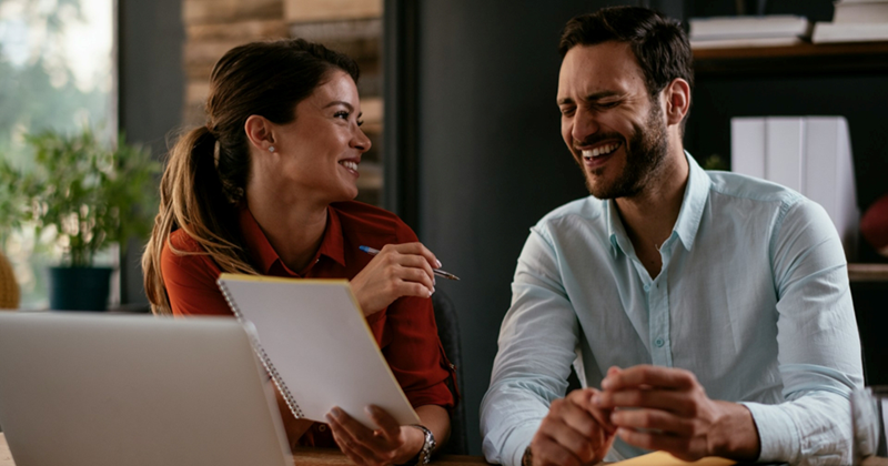 A woman and a man sitting in front of a laptop laugh together as the woman holds a notepad
