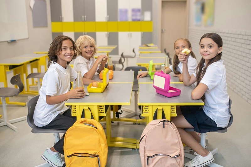 A group of young kids sitting down at a school table wearing uniforms eating lunch.