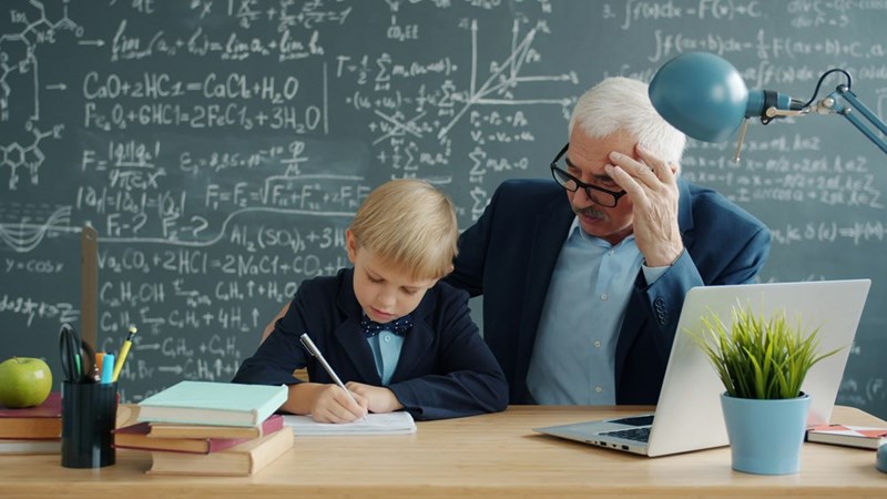Old man tutoring a kid in a classroom wearing a suit, writing and reading.