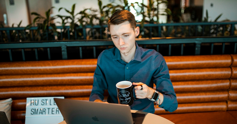 A man sits and works on a MacBook while holding a mug