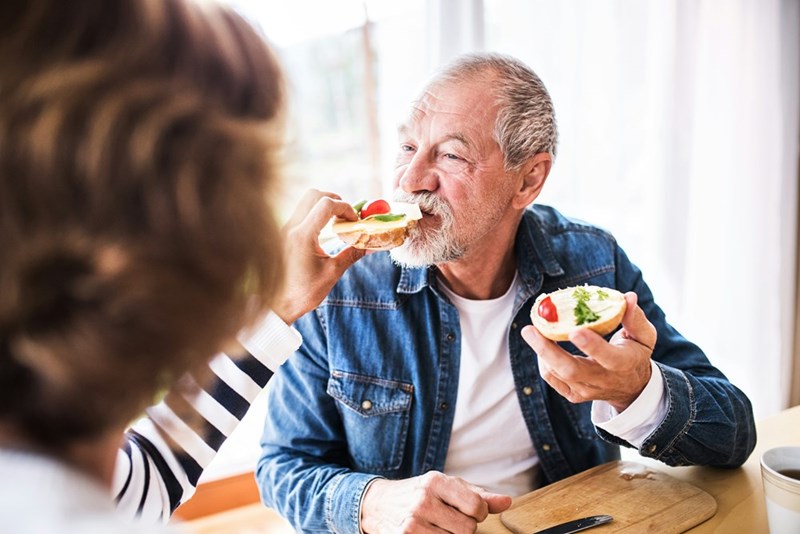 Family members helping a grandpa eat a sandwich.