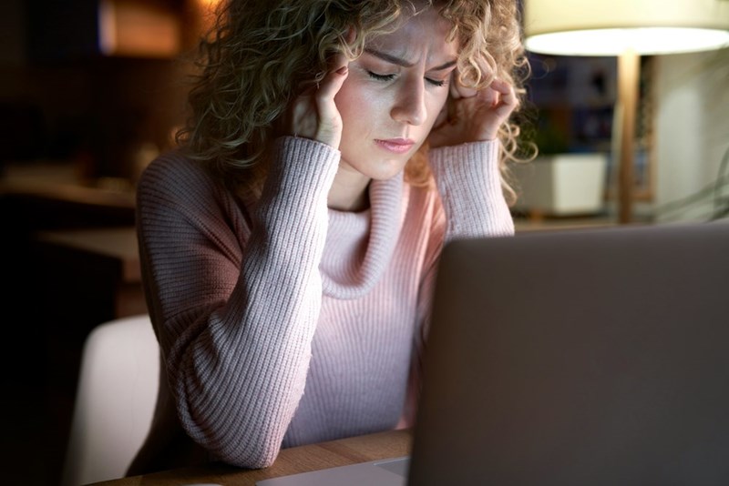 Stressed woman using a laptop at night while holding her temples in a dimly lit room.