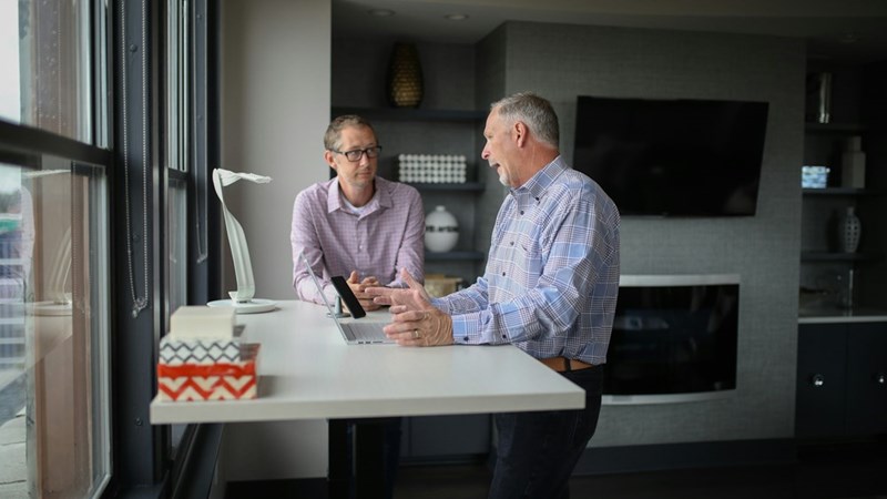 Two men talking during a meeting at a standing desk in a modern office with laptops and large windows.
