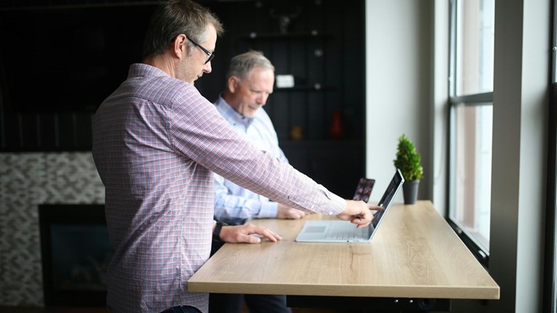 Two coworkers reviewing a laptop together at a standing desk in a modern office with natural window light.