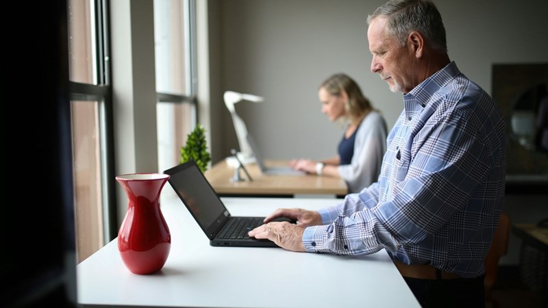 Older man using a laptop at a standing desk near office windows with a coworker working in the background.
