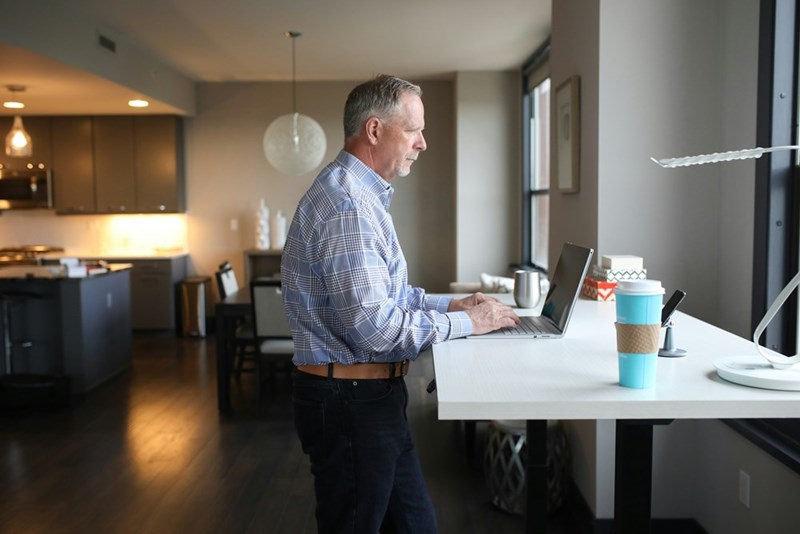 Older man using a laptop at a standing desk in a modern apartment home office beside large windows.