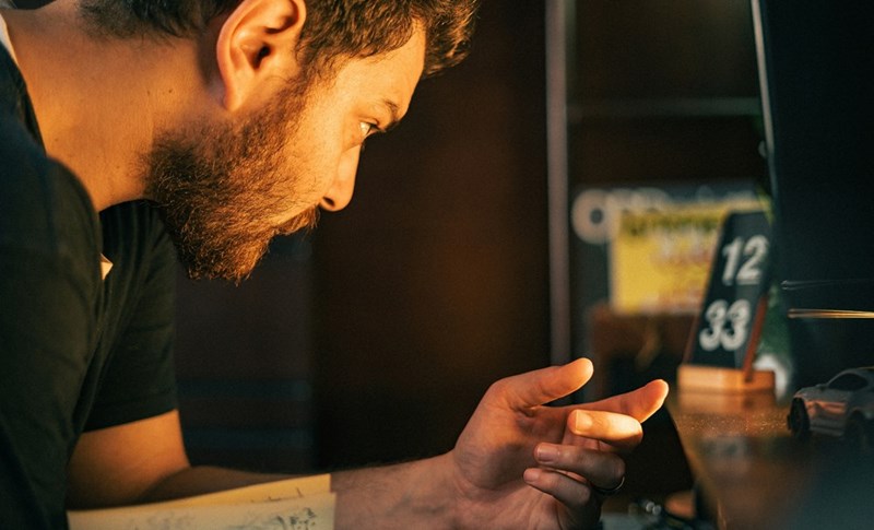 Man checking his smartphone late at night beside a digital clock and coffee mug in warm indoor lighting.