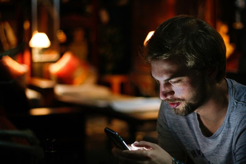Young man checking his smartphone at night in a dimly lit room with warm ambient lighting behind him.