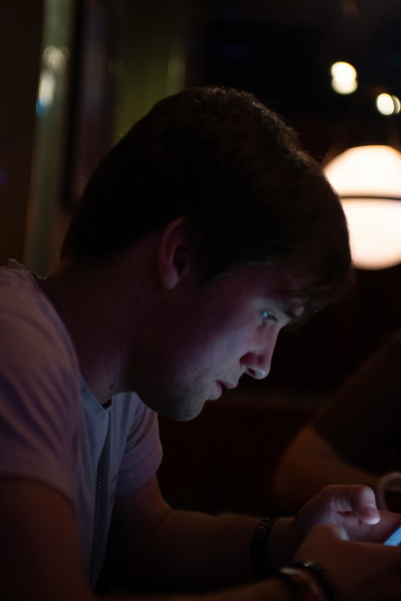 Young man using a smartphone in a dark room with screen light illuminating his face at night.