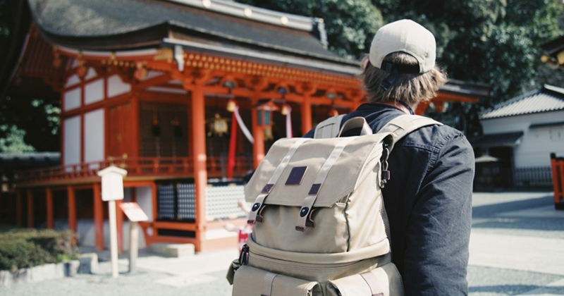 The back of a man wearing a cap and a backpack, walking past a Japanese temple
