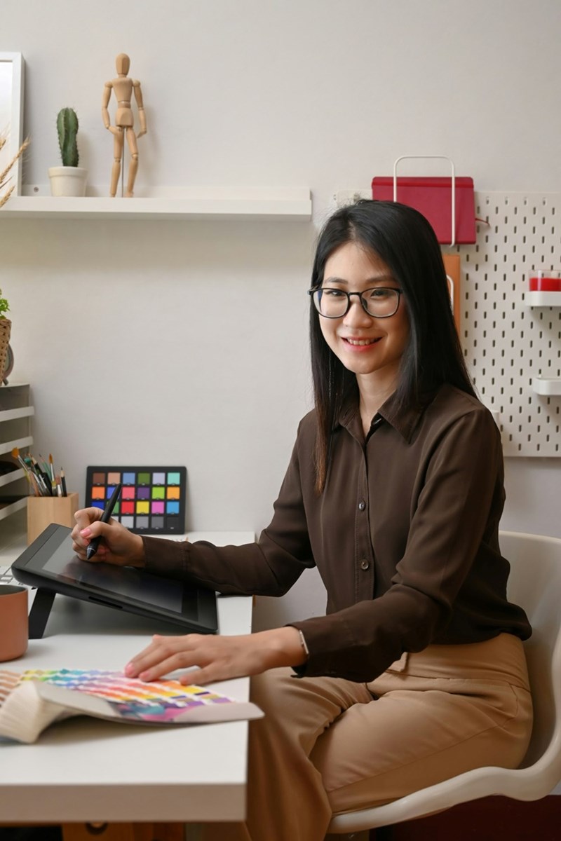 A woman sits at a desk with a drawing tablet and a color palette